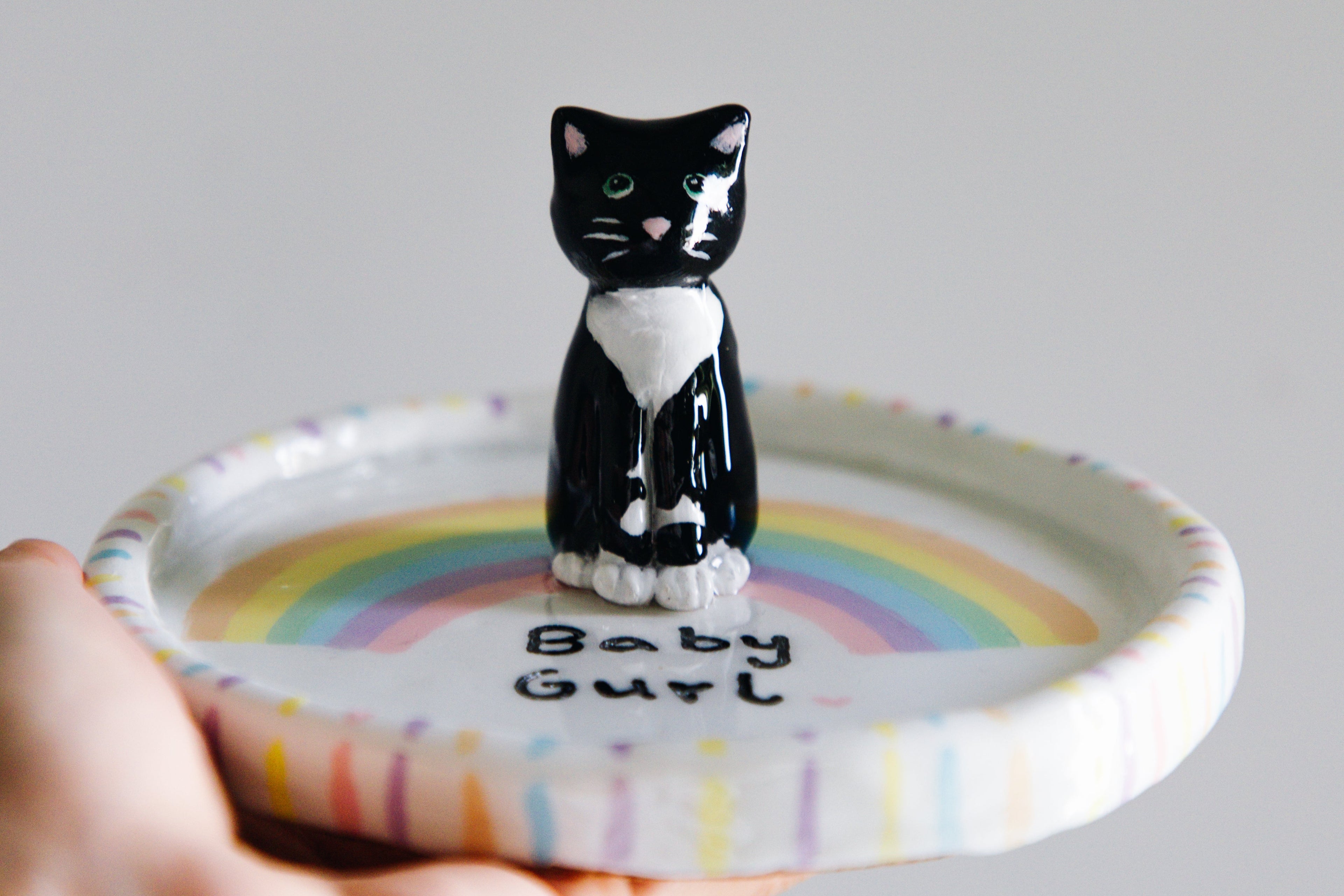 Person holding a small round tray with a black and white cat figurine and 'Baby Girl' text on a gray background